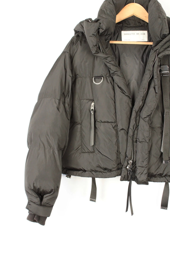 Shoreditch Ski Club black cropped puffer jacket with quilted matte shell and harness straps.